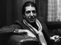 Leonard Cohen, mit Schal und Zigarette, sitzt nachdenklich auf einem Ledersofa. Ein ikonisches Bild, das den Geist des Sängers einfängt.