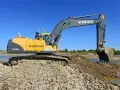 Żółta koparka Volvo EC240B LC pracuje na kamienistym brzegu rzeki. Zastanawiasz się, ile kosztuje godzina pracy koparki?