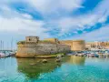 A stone castle overlooks a harbor filled with boats and yachts under a bright blue sky.