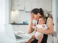 Zmęczona mama z płaczącym niemowlakiem na rękach, próbuje pracować przy laptopie. Czuje przebodźcowanie niemowlaka i własne.