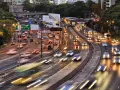 Traffico intenso su un'autostrada urbana al tramonto. Le luci dei veicoli creano scie luminose, con un **segnale 105** che indica la direzione.