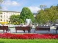 Park z fontanną i ludźmi siedzącymi na ławkach, w tle budynek.