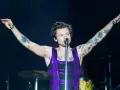 Harry Styles w błyszczącym fioletowym topie śpiewa na koncercie, unosząc ramiona. Jego ramiona pokryte są tatuażami.