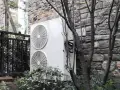 Dwa wentylatory jednostki zewnętrznej klimatyzatora sprsun, zamontowane na kamiennej ścianie, otoczone zielenią i gałęziami drzew.