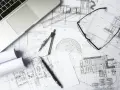 Na biurku architekta leżą plany budowlane, laptop, okulary i cyrkiel. Trwają prace nad przedmiarem robót.