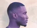 Perfil de hombre con taper fade medio texturizado, barba corta y cadena. Fondo abstracto con destellos.