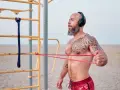 Man med tatueringar och skägg tränar med gummisnoddar vid ett utegym på stranden.