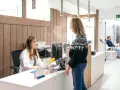 Uśmiechnięta rejestratorka medyczna w białym fartuchu obsługuje pacjentkę. Czy to zawód medyczny? W poczekalni siedzi mężczyzna.