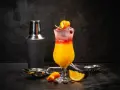 Kolorowy drink z lodem, pomarańczą i żurawiną, obok shakera. Wybór między Cosmopolitan a Sex on the Beach.