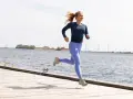 En kvinna springer glatt på en träbrygga vid havet, med vindkraftverk i bakgrunden. Hon verkar njuta av att springa 1 km varje dag.