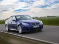 Niebieskie BMW M5 E60, ikona designu Chrisa Bangle'a, pędzi po drodze wśród zielonych pól i błękitnego nieba.