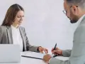 Kobieta i mężczyzna w garniturach przy biurku, podpisują dokumenty.