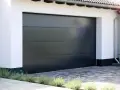 En modern, mörkgrå garageport montering med horisontella linjer. En vit fasad och grå stenläggning framför.
