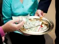 A hand places a twenty-dollar bill onto a golden tray filled with cash and coins, a gesture of offering to the poor.