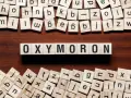 Holzbuchstaben bilden das Wort "OXYMORON". Sie sind umgeben von weiteren Buchstabenkacheln, die an Spiele wie Scrabble erinnern und Beispiele für Oxymoron-Begriffe darstellen könnten.