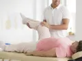Ein Physiotherapeut unterstützt eine ältere Frau bei einer Beinübung im Rahmen einer PNF Therapie.