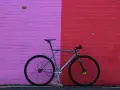 A sleek, fixed gear bike with "MASH" branding leans against a vibrant pink and red brick wall.