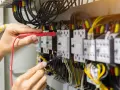 Elektryk sprawdza przewód neutralny w szafie sterowniczej, używając multimetru. Widać wiele kabli i przekaźników.
