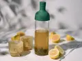 Orzeźwiający cold brew tea w butelce z zieloną zakrętką, obok szklanki z lodem i cytryny. Idealny na upalne dni.