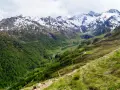 Un sentiero serpeggia su un pendio erboso verso un passo rombo in moto, con montagne innevate sullo sfondo.