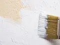 Un pinceau applique un badigeon de chaux blanc sur un mur texturé de couleur beige.