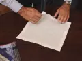 Mani esperte mostrano come piegare fazzoletto da taschino su un tavolo di legno. Altri fazzoletti colorati sono visibili a sinistra.