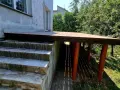 Nowy, wysoki taras na słupach, z drewnianym blatem, sąsiaduje z betonowymi schodami i zielonym ogrodem.
