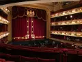 Ein prunkvolles Opernhaus mit roten Samtvorhängen und goldenen Verzierungen. Die Logen sind mit Lichtern geschmückt, und im Parkett sitzen Zuschauer.