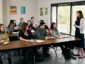 Uczniowie słuchają nauczycielki podczas lekcji. W klasie panuje atmosfera skupienia, jak podczas ważnej rady pedagogicznej.