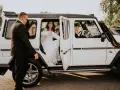 Uśmiechnięta panna młoda w sukni ślubnej wysiada z białego samochodu do ślubu. Obok stoi mężczyzna w garniturze.