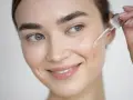 Una mujer sonríe mientras aplica un suero con antioxidantes faciales en su piel con un gotero.