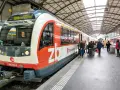 A train labeled "PE Interlaken, Ost" waits at the station, ready for its journey on the Lucerne to Interlaken Golden Pass line. Passengers gather on the platform.