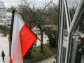 Flaga państwowa w biało-czerwonych barwach powiewa na wietrze. W tle widać drzewa i budynki.