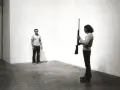 Chris Burden, dans une performance tendue, tient un fusil. Un homme se tient à distance, le regard fixé.
