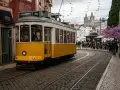 Żółty tramwaj 28 na brukowanej ulicy w Lizbonie, z ludźmi czekającymi na przystanku i kościołem w tle.