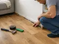 Hombre arreglando tarima flotante que cruje al pisar. Martillo, cinta y cuña verde listos para la solución.