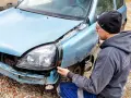 Mężczyzna w czapce i bluzie naprawia niebieską ćwiartkę samochodu, która ucierpiała w wypadku.