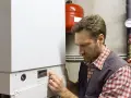 Un tecnico con una camicia a quadri e un gilet grigio regola un pannello di controllo di una caldaia bianca. Guida nella scelta della caldaia.