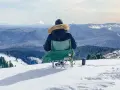 Persona sentada en silla plegable en la nieve, mirando un paisaje montañoso cubierto de árboles.