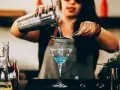Barman przygotowuje drinka, przelewając niebieski płyn przez sitko do kieliszka. W tle widać butelki z różnymi nazwami drinków alkoholowych.