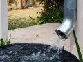 Woda z rynny spływa do czarnej beczki, zbierając deszczówkę. W tle widać drewniany taras i jesienny ogród.