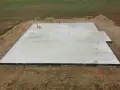 Płyta fundamentowa koszt budowy domu. Betonowa płyta fundamentowa z wystającymi rurami i elementami instalacji, otoczona ziemią i żwirem.