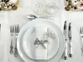 Ett elegant dukat bord med vita tallrikar, servetter dekorerade med blommor och vackra, silverfärgade engångsbestick.