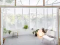 Interno luminoso con piante, sedia bianca e sgabello in legno, affacciato su una strada alberata da una veranda balcone.