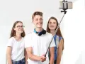 Trzech przyjaciół robi selfie na kijku. Chłopak z przodu uśmiecha się szeroko, dziewczyna w okularach po lewej stronie też, a ta z tyłu patrzy w obiektyw. To świetny sposób, jak robic dobre zdjecia!