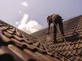 Pracownik w kasku i uprzęży zabezpieczającej wykonuje renowację dachu, natryskując dachówkę.