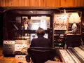 Osoba siedzi przed komputerem w profesjonalnym **recording studio**, otoczona sprzętem audio i mikserem.