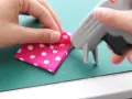Mani che piegano un tessuto rosa a pois bianchi mentre una pistola per colla a caldo deposita un punto. La colla a caldo si stacca con l'acqua, un dettaglio importante per i lavoretti.