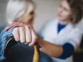 Opiekunka wspiera starszą panią, która trzyma laskę. Zastanawiasz się, ile kosztuje opiekunka z mops?