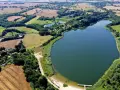 Malownicze jezioro Marszewo otoczone zielenią i polami. Widać piaszczystą plażę z pomostem i kilka samochodów.